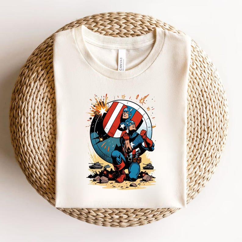 Puede incluir: Camiseta de color crema con un gr&aacute;fico del Capit&aacute;n Am&eacute;rica. El dise&ntilde;o muestra al Capit&aacute;n Am&eacute;rica arrodillado con su escudo, un escudo rojo, blanco y azul, y explosiones. La camiseta est&aacute; doblada sobre una superficie circular tejida.