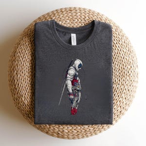 Puede incluir: Una camiseta gris marengo de cuello redondo con una ilustraci&oacute;n gr&aacute;fica de un personaje con m&aacute;scara blanca, equipo t&aacute;ctico rojo y azul, y una espada en la espalda. El personaje aparece de perfil sobre un fondo circular tenue y sutil.