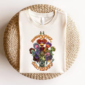 May include: A cream-colored crew neck t-shirt featuring a colorful graphic of Fraggle Rock characters. The design includes the text "Fraggle Rock 30th Anniversary" in orange lettering, surrounding a group of various puppet characters in vibrant colors.