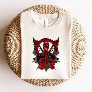 K&ouml;nnte beinhalten: Cremefarbenes T-Shirt mit einer Grafik von Deadpool in Gebetshaltung. Das Design umfasst eine rote Deadpool-Maske in einem roten Kreis, zwei gekreuzte Schwerter sowie schwarze und rote Akzente. Das Shirt wird auf einer gewebten Oberfl&auml;che pr&auml;sentiert.