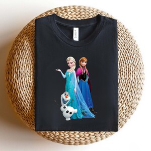 Puede incluir: Camiseta negra con Elsa, Anna y Olaf de la pel&iacute;cula Frozen. Elsa lleva un vestido azul con una larga capa fluida. Anna viste un vestido azul con una capa rosa. Olaf es un mu&ntilde;eco de nieve blanco con nariz de zanahoria y brazos de ramitas.