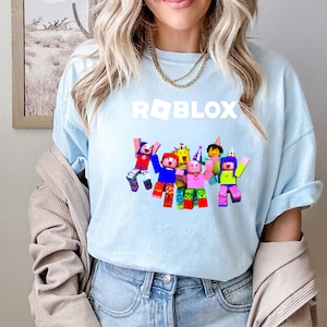 May include: Light blue t-shirt featuring the word "ROBLOX" in white block letters above a graphic of several blocky characters with party hats and raised arms. The characters are in various colors, including red, blue, yellow, pink, and green.