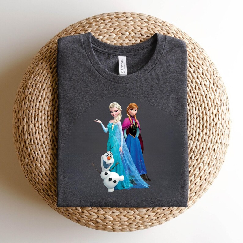 Puede incluir: Camiseta gris oscuro con Elsa, Anna y Olaf de la pel&iacute;cula Frozen. Elsa lleva un vestido azul, Anna un vestido y capa azules, y Olaf es un mu&ntilde;eco de nieve blanco. La camiseta es de un material suave.