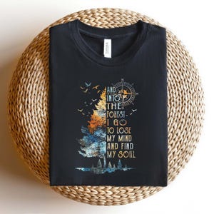 May include: A black crew neck t-shirt featuring a graphic print of a split-colored tree in orange and blue, a compass rose, and birds in flight. The text reads: AND INTO THE FOREST I GO TO LOSE MY MIND AND FIND MY SOUL in a stylized serif font.