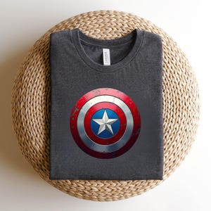 May include: A dark charcoal grey crew neck t-shirt featuring a centred graphic of a circular shield with a distressed red, white, and blue finish and a central white star. The shirt has a white brand label visible at the inner neckline.