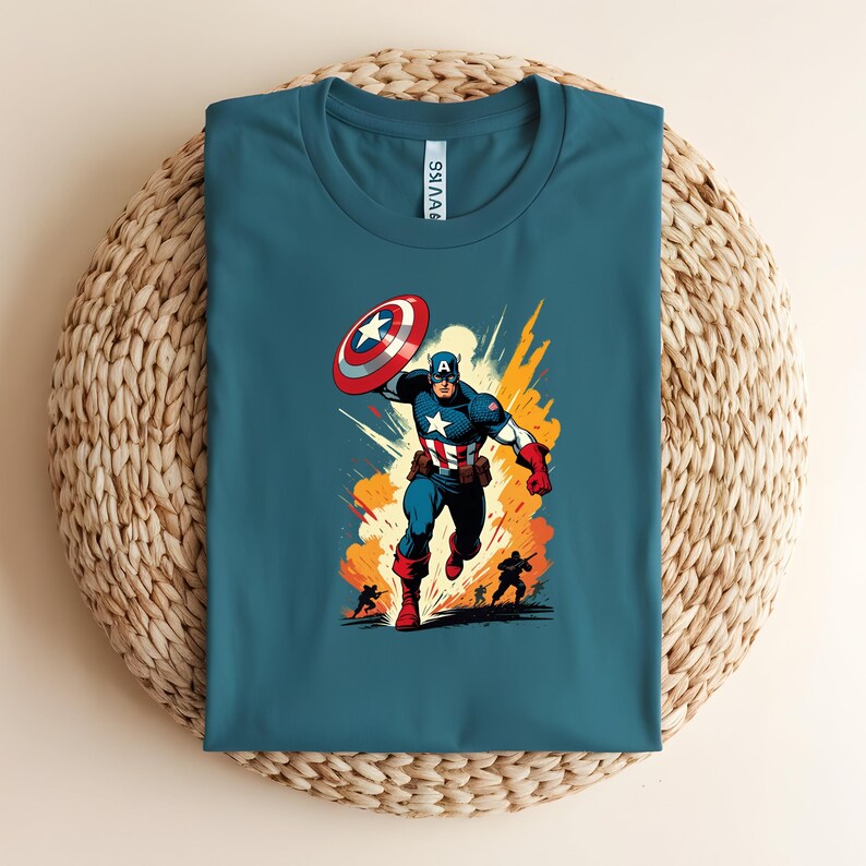 May include: Teal crew neck t-shirt featuring a comic-style illustration of Captain America. The graphic shows Captain America in action, holding his shield, with a fiery background. The shirt is displayed on a woven, circular surface.