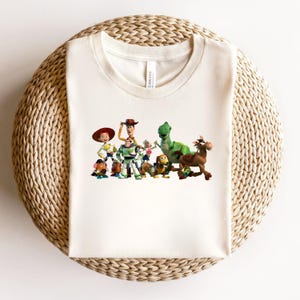 May include: Cream-colored t-shirt featuring a colorful graphic of characters from the animated film Toy Story. The graphic includes Woody, Buzz Lightyear, Rex, and other characters. The shirt is made of a soft material and is displayed on a woven surface.