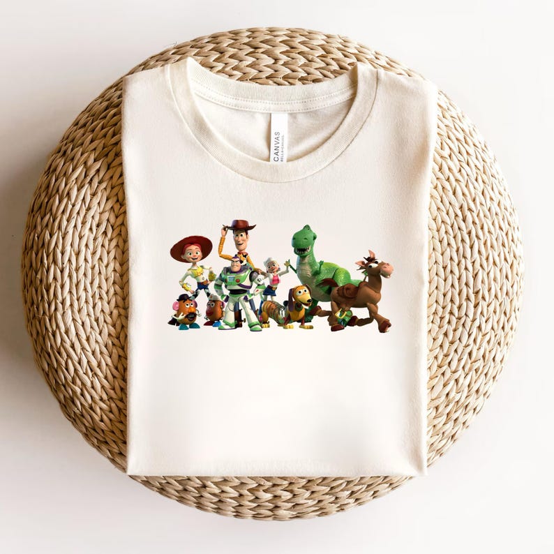 May include: Cream-colored t-shirt featuring a colorful graphic of characters from the animated film Toy Story. The graphic includes Woody, Buzz Lightyear, Rex, and other characters. The shirt is made of a soft material and is displayed on a woven surface.