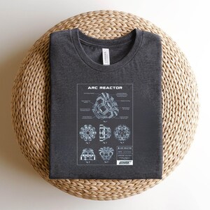 Puede incluir: Camiseta gris oscuro con un diagrama del Reactor Arc, con el texto "ARC REACTOR". El diagrama muestra esquemas detallados y el logotipo de Stark Industries. La camiseta se muestra sobre una superficie tejida.