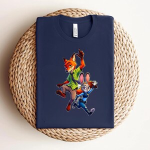 Puede incluir: Una camiseta azul marino de manga corta con un estampado gr&aacute;fico de un zorro de dibujos animados con camisa verde y corbata, junto a un conejo de dibujos animados con uniforme de polic&iacute;a azul. La camiseta tiene una etiqueta blanca en el cuello.