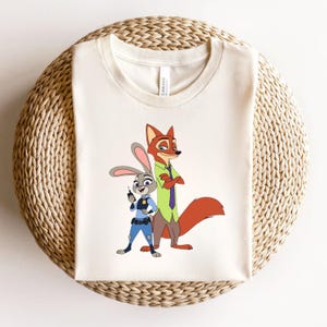Peut inclure: Un t-shirt &agrave; manches courtes de couleur cr&egrave;me avec une impression graphique repr&eacute;sentant un lapin policier en uniforme bleu et un renard portant une chemise vert clair avec une cravate violette, debout c&ocirc;te &agrave; c&ocirc;te sur le devant du v&ecirc;tement.