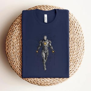 Op de afbeelding: Marineblauw t-shirt met een gedetailleerde illustratie van Iron Man. Het ontwerp toont het pantser van het personage met een gloeiende boogreactor. Het shirt is op een geweven oppervlak gevouwen.
