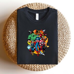 May include: A black crew neck t-shirt featuring a colorful graphic print of various comic book superheroes in a space setting with a planet in the background. The shirt has a white tag at the inner neckline with the brand name Canvas printed on it.