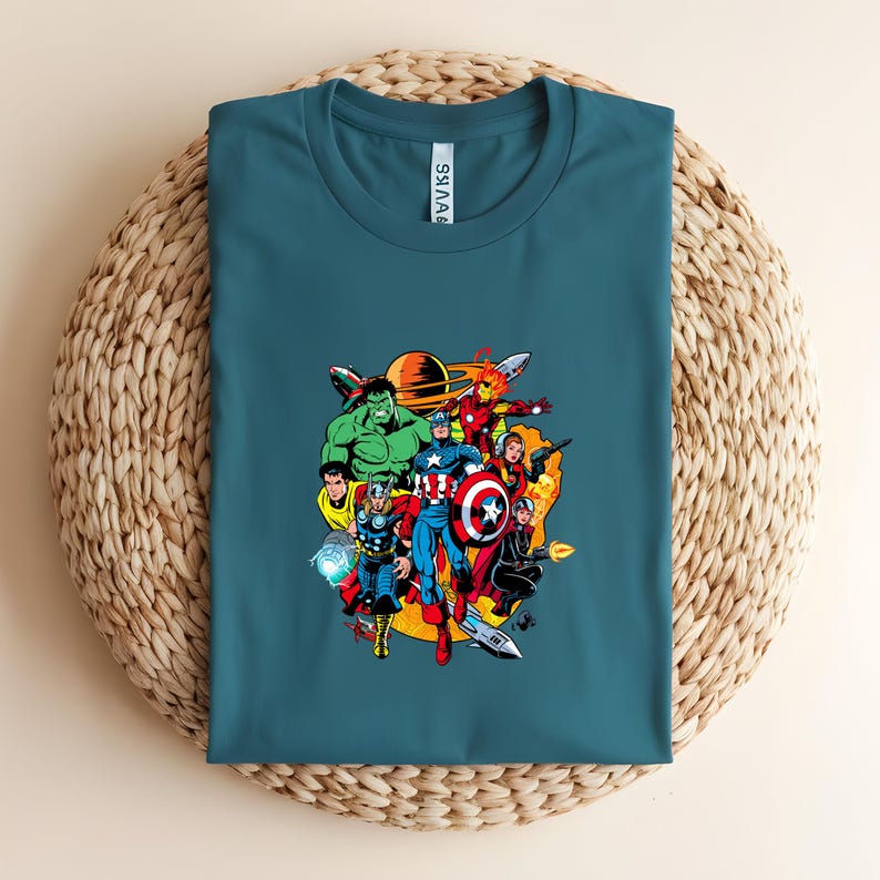 May include: A teal crew neck t-shirt featuring a colorful graphic print of various comic book superheroes including Hulk, Thor, and Captain America. The design includes space elements like a planet and rockets, with the text "AVKS" on the neck label.