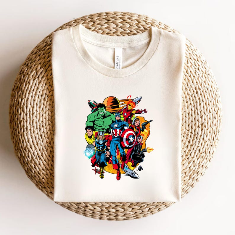 May include: A cream-colored crew neck t-shirt featuring a colorful graphic print of various comic book superheroes, including Hulk, Thor, Captain America, and Iron Man, arranged in a dynamic cluster against a yellow circular background.