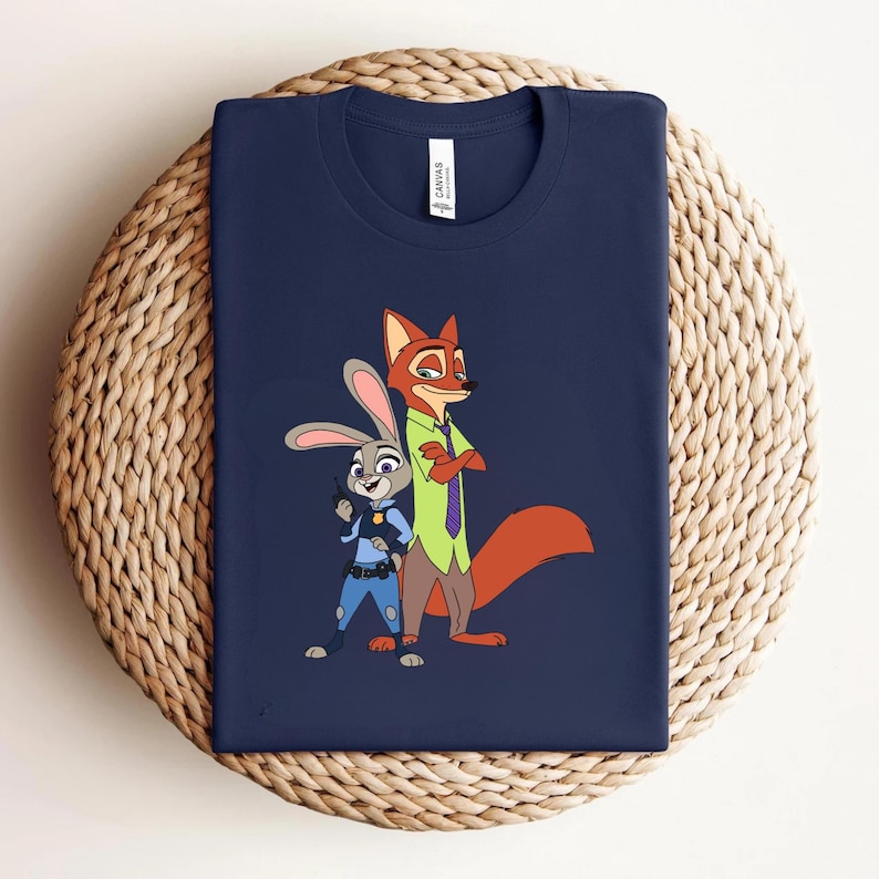 Peut inclure: Un t-shirt bleu marine &agrave; col rond avec une impression graphique repr&eacute;sentant un lapin de dessin anim&eacute; en uniforme de police bleu et un renard de dessin anim&eacute; en chemise vert clair et cravate violette. Une &eacute;tiquette blanche est visible au col.