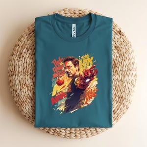 May include: Teal t-shirt featuring a colorful graphic of Iron Man with the text "The Tone OK" and other phrases. The artwork depicts Iron Man pointing with his armored hand. The shirt is folded on a woven surface.
