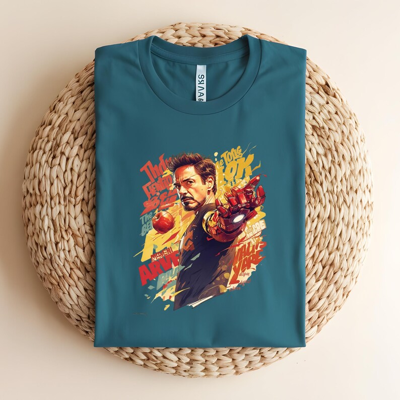 May include: Teal t-shirt featuring a colorful graphic of Iron Man with the text "The Tone OK" and other phrases. The artwork depicts Iron Man pointing with his armored hand. The shirt is folded on a woven surface.