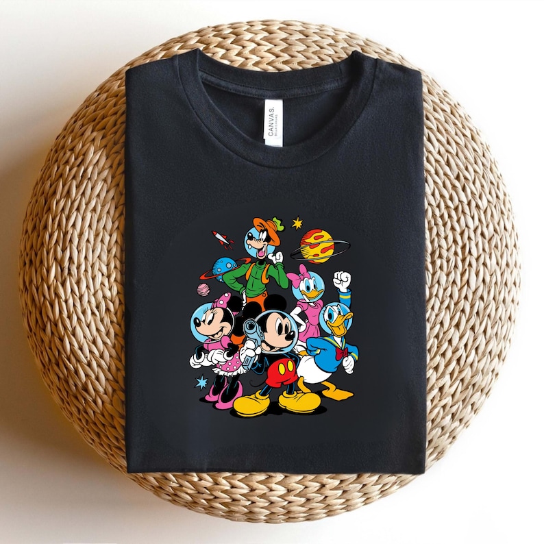 May include: Black t-shirt featuring a colorful graphic of Mickey Mouse, Minnie Mouse, Donald Duck, Daisy Duck, and Goofy in space suits. The cartoon characters are surrounded by planets, stars, and a rocket ship.