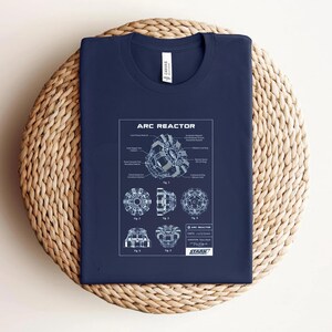 Puede incluir: Camiseta azul marino con un esquema blanco de un Reactor Arc, con diagramas y etiquetas detalladas. El texto "ARC REACTOR" est&aacute; en la parte superior, con "STARK" en la parte inferior. La camiseta se muestra sobre una superficie circular tejida.