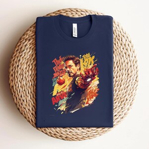 May include: Navy blue t-shirt featuring a colorful graphic of Iron Man, with the actor's likeness and text elements. The design includes red, yellow, and orange accents. The shirt is folded on a woven, circular surface.
