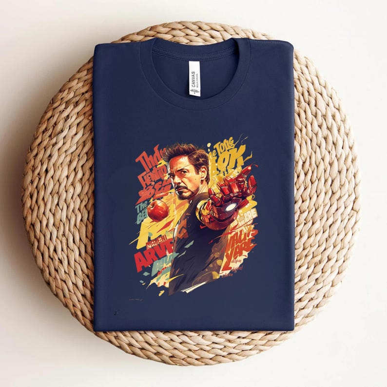 May include: Navy blue t-shirt featuring a colorful graphic of Iron Man, with the actor's likeness and text elements. The design includes red, yellow, and orange accents. The shirt is folded on a woven, circular surface.