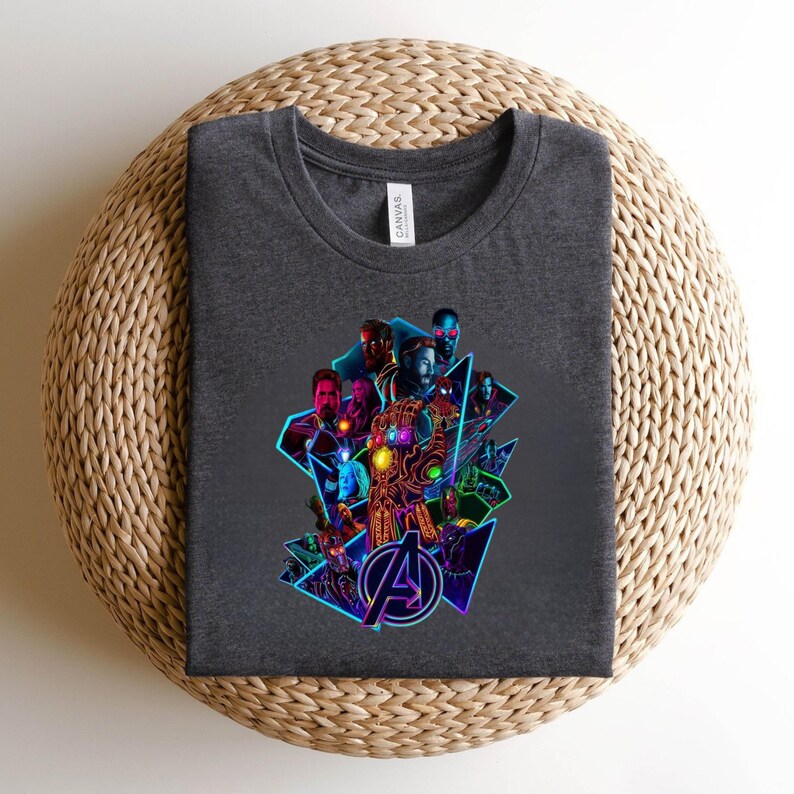 May include: Dark grey t-shirt with a vibrant graphic of several superheroes and the Avengers logo. The design incorporates geometric shapes and neon blue and purple highlights. The tee is made of a soft fabric.