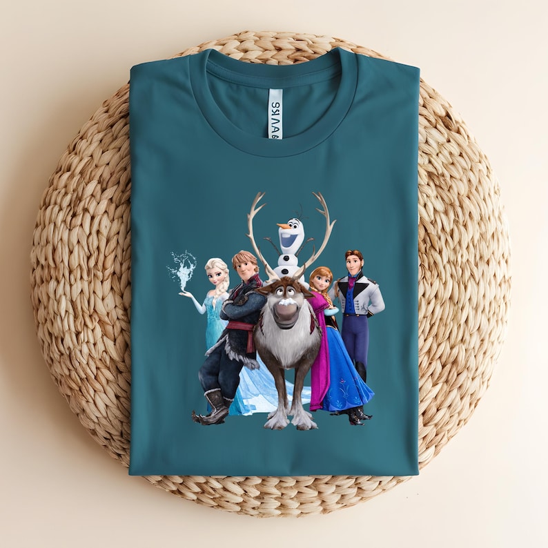 Op de afbeelding: Teal t-shirt met een afbeelding van personages uit de animatiefilm "Frozen". Het ontwerp bevat Elsa, Anna, Kristoff, Olaf en Sven. Het shirt is opgevouwen op een geweven, cirkelvormig oppervlak.