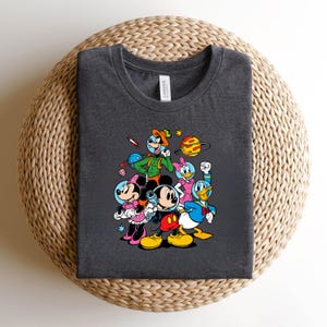 May include: Dark gray t-shirt featuring a colorful graphic of Mickey Mouse, Minnie Mouse, Donald Duck, Daisy Duck, and Goofy in space suits, with planets and stars. The shirt is made of a soft material and has a crew neck.
