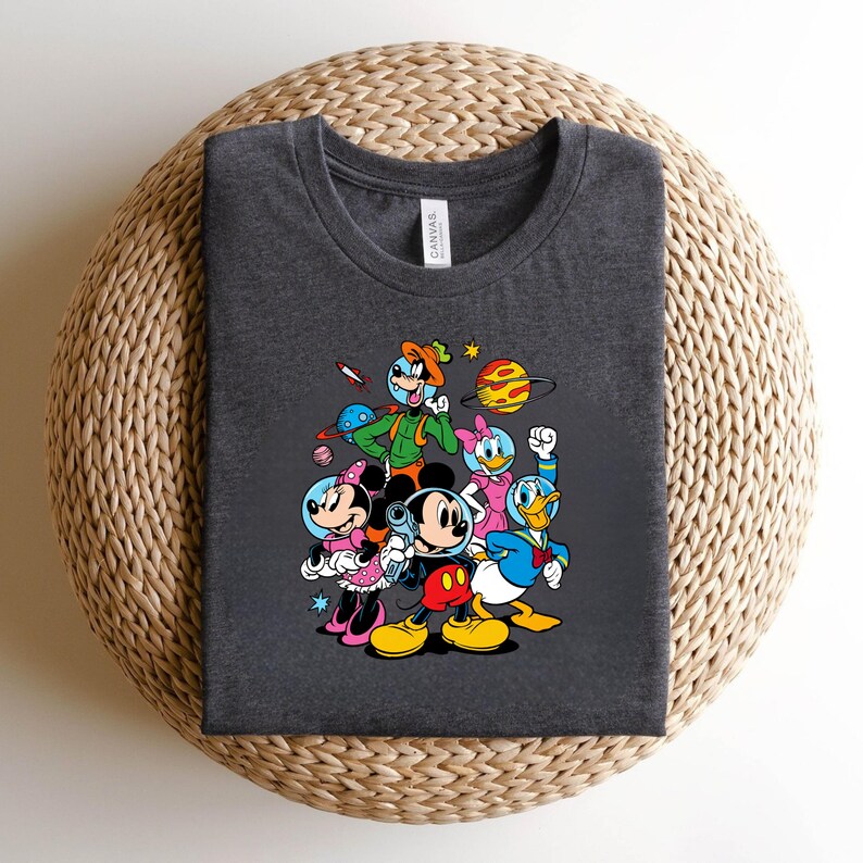 May include: Dark gray t-shirt featuring a colorful graphic of Mickey Mouse, Minnie Mouse, Donald Duck, Daisy Duck, and Goofy in space suits, with planets and stars. The shirt is made of a soft material and has a crew neck.