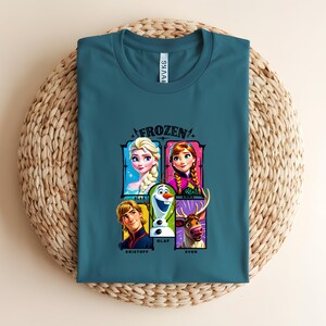 May include: Teal t-shirt featuring the word "FROZEN" above a graphic of characters Elsa, Anna, Olaf, Kristoff, and Sven. The shirt is folded on a woven, circular surface.