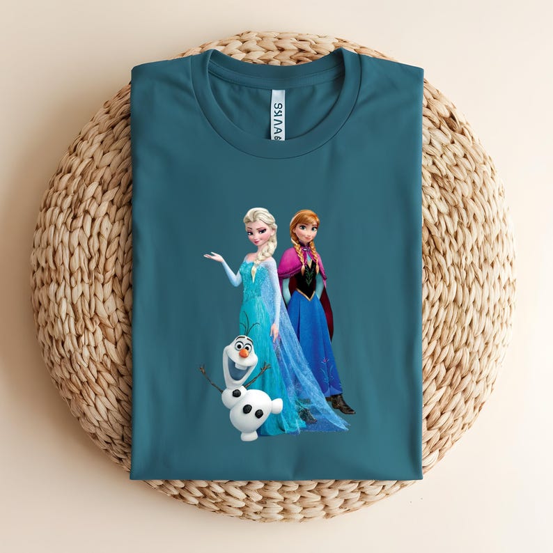 Puede incluir: Camiseta verde azulado con Elsa, Anna y Olaf de Frozen. Elsa lleva un vestido azul, Anna un vestido azul y morado, y Olaf es un mu&ntilde;eco de nieve blanco. La camiseta est&aacute; doblada sobre una superficie tejida.