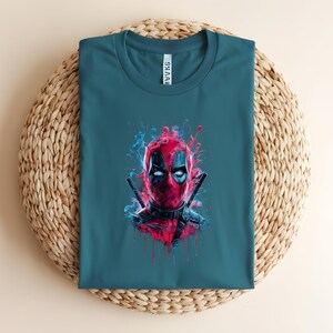 May include: Teal t-shirt featuring a vibrant graphic of Deadpool's face. The artwork showcases Deadpool's mask with red and black details, surrounded by blue and red artistic smoke effects. Two katanas are crossed behind the head.