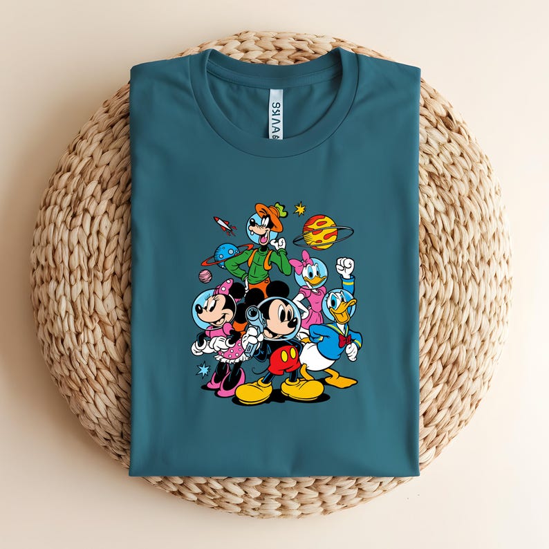 May include: Teal crew neck t-shirt featuring a colorful graphic of cartoon characters in space suits. The graphic includes Mickey Mouse, Minnie Mouse, Donald Duck, and other Disney characters with planets and a rocket ship.