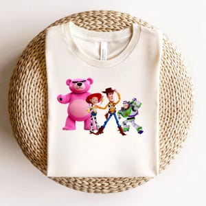 May include: Off-white t-shirt featuring a colorful graphic of characters from the animated film Toy Story. The design includes a pink bear, Jessie, Woody, and Buzz Lightyear. The shirt is made of a soft material and is displayed on a woven surface.