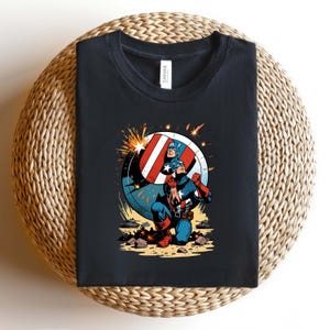 Puede incluir: Camiseta negra con un gr&aacute;fico de Capit&aacute;n Am&eacute;rica en acci&oacute;n. La ilustraci&oacute;n muestra al Capit&aacute;n Am&eacute;rica arrodillado con su escudo. El escudo es rojo, blanco y azul, con una estrella. El dise&ntilde;o es de estilo c&oacute;mic.