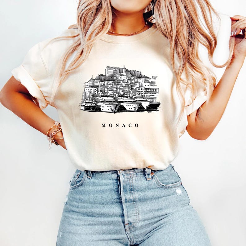 K&ouml;nnte beinhalten: Cremefarbenes T-Shirt mit einer Schwarz-Wei&szlig;-Illustration der Skyline von Monaco und Yachten. Das Wort "MONACO" ist darunter gedruckt. Das Shirt wird mit einer blauen Jeans kombiniert.