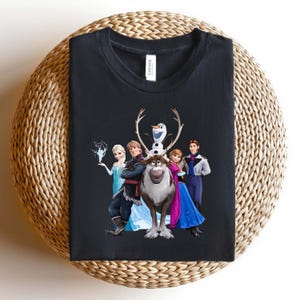 Op de afbeelding: Zwart T-shirt met een kleurrijke afbeelding van personages uit de animatiefilm Frozen. Het ontwerp omvat Elsa, Anna, Kristoff, Olaf en Sven. Het shirt is op een geweven oppervlak gevouwen.