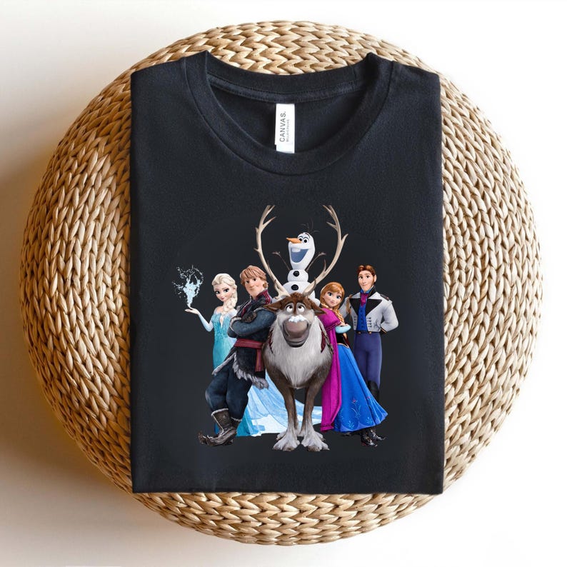 Op de afbeelding: Zwart T-shirt met een kleurrijke afbeelding van personages uit de animatiefilm Frozen. Het ontwerp omvat Elsa, Anna, Kristoff, Olaf en Sven. Het shirt is op een geweven oppervlak gevouwen.