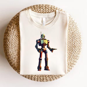 May include: Cream-colored t-shirt featuring a colorful robot illustration. The robot has a green, yellow, and black head, with a matching body and legs. It is holding a weapon in its right hand. The shirt is made of a soft material.