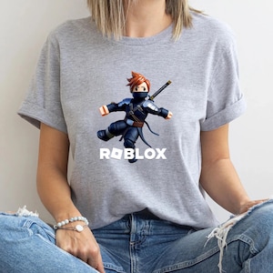 May include: A gray t-shirt featuring a Roblox ninja character in a dynamic pose. The ninja is clad in a dark blue outfit with a black mask and a sword. The Roblox logo is printed in white below the character.