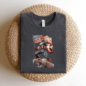 Pode incluir: T-shirt cinza escuro com um gr&aacute;fico do Capit&atilde;o Am&eacute;rica segurando uma bandeira americana e um escudo. O design inclui o ic&ocirc;nico escudo estrelado e as cores vermelha, branca e azul da bandeira. A camisa &eacute; feita de um material macio.