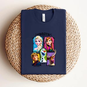 May include: Navy blue t-shirt featuring a colorful graphic of characters from the movie "Frozen." The design includes Elsa, Anna, Olaf, Kristoff, and Sven. The word "FROZEN" is printed above the characters.