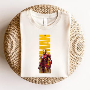May include: Cream-colored t-shirt featuring a graphic of Iron Man in his red and gold armor. The word "MAN" is printed vertically in yellow, with "IRON" above it. The shirt is made of a soft material and is laid on a woven surface.