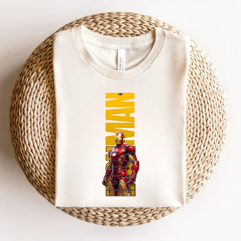 May include: Cream-colored t-shirt featuring a graphic of Iron Man in his red and gold armor. The word "MAN" is printed vertically in yellow, with "IRON" above it. The shirt is made of a soft material and is laid on a woven surface.