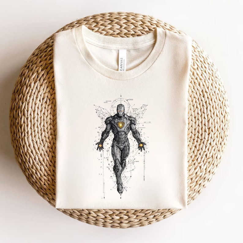Op de afbeelding: Cr&egrave;mekleurig T-shirt met een gedetailleerde illustratie van een superheld in vliegende pose. Het ontwerp is voornamelijk zwart-wit met gouden accenten op de borst en handen. Het shirt is op een geweven, cirkelvormig oppervlak gevouwen.