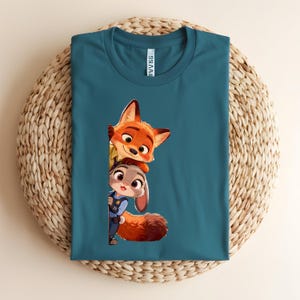 May include: A teal crew neck t-shirt featuring a graphic print of a cartoon fox and a cartoon rabbit peeking out from the side. The fox has orange fur and the rabbit has gray fur and is wearing a blue uniform. The shirt has a white neck label.