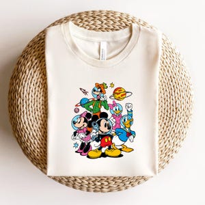 May include: Off-white t-shirt featuring a colorful graphic of cartoon characters in space suits. Mickey Mouse, Minnie Mouse, Donald Duck, and Daisy Duck are among the characters. A rocket ship and planets are also included in the design.