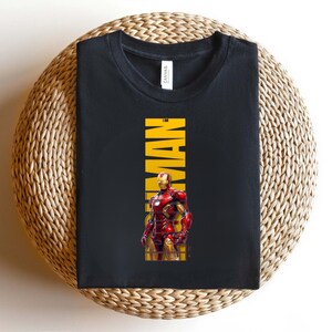 May include: Black t-shirt featuring a graphic of Iron Man in his red and gold armor. The word "IRONMAN" is printed vertically in yellow. The shirt is folded on a woven, circular surface.