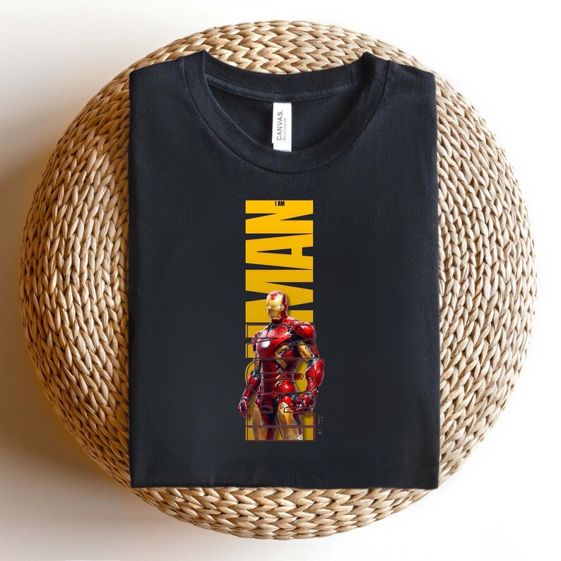 May include: Black t-shirt featuring a graphic of Iron Man in his red and gold armor. The word "IRONMAN" is printed vertically in yellow. The shirt is folded on a woven, circular surface.